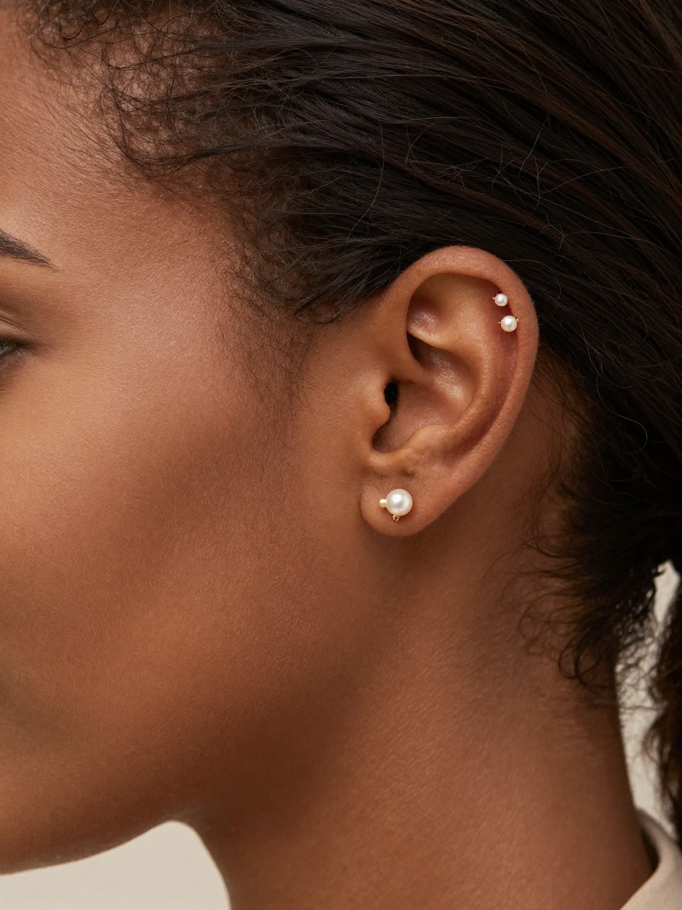 Side portrait close-up of an ear with deep brown skin tone, hair tucked neatly behind the ear, three tiny pearl studs placed along the upper helix in a clean rising line, soft gold accents, realistic skin texture, natural daylight, shallow depth of field, elegant editorial beauty photography, focus fully on the upper ear and earrings.