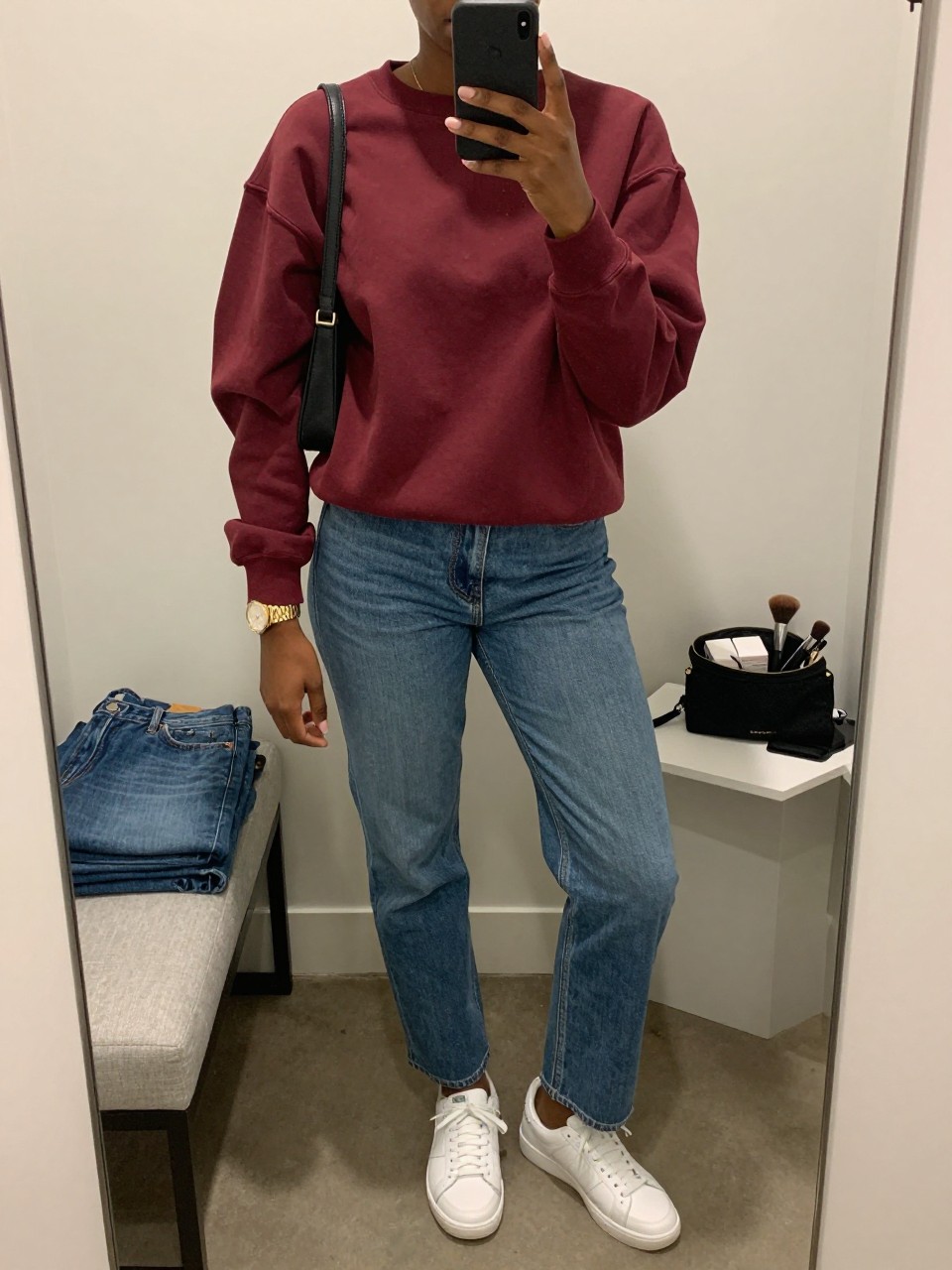 mirror selfie of a Black woman wearing the exact outfit described above, clearly showing a burgundy oversized crew neck sweatshirt, medium wash relaxed straight-leg jeans, white leather sneakers, black shoulder bag, and gold watch, holding a phone in front of a mirror, cropped at NECK so the face does not show, with a relaxed casual selfie pose, in a walk-in closet mirror, soft indoor lighting, including folded denim jeans on a bench and a makeup brush or cosmetic bag on the counter, latest iPhone photo quality.