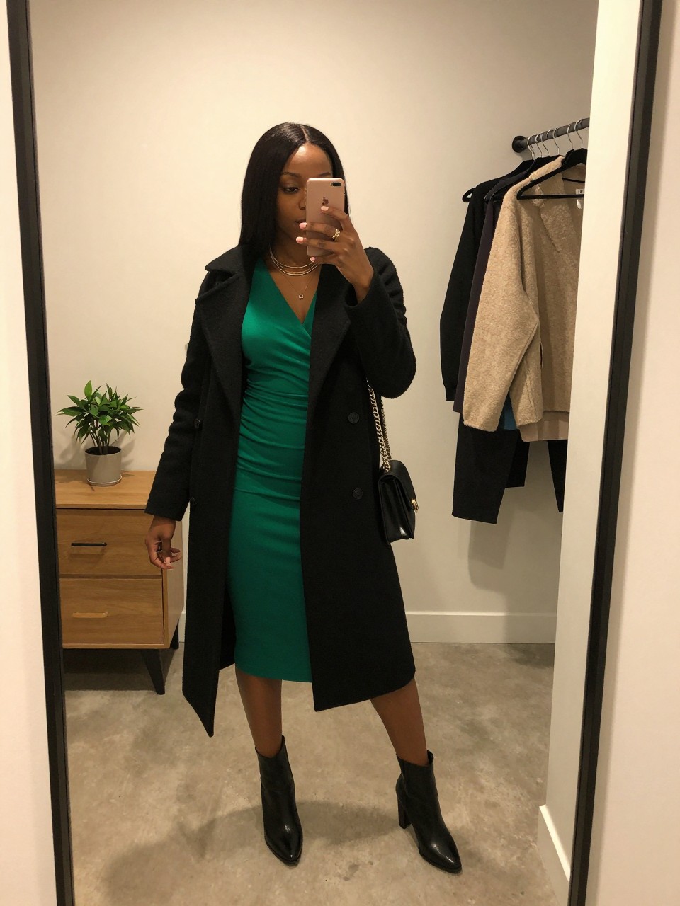 mirror selfie of a Black woman wearing the exact outfit described above, clearly showing an emerald green long-sleeve wrap dress, black heeled ankle boots, black wool-blend coat, black chain shoulder bag, and gold layered necklace, holding a phone in front of a mirror, cropped at NECK so the face does not show, with a relaxed birthday selfie pose, in a minimal dressing room, warm moody lighting, including a small plant on a dresser and a clothing rack with garments behind, latest iPhone photo quality.