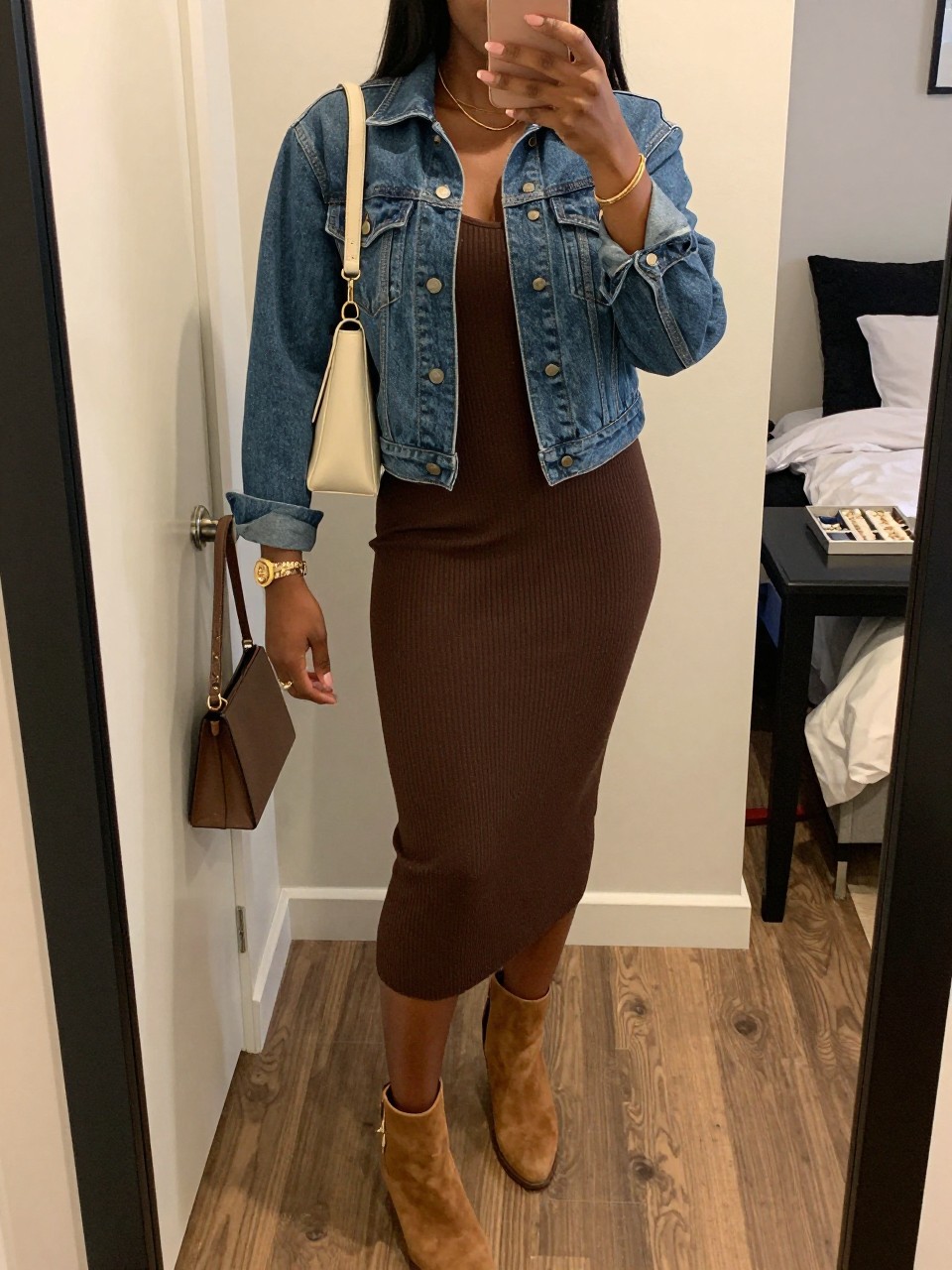 mirror selfie of a Black woman wearing the exact outfit described above, clearly showing a medium wash denim jacket, chocolate brown ribbed knit midi dress, tan ankle boots, cream shoulder bag, and gold bracelet, holding a phone in front of a mirror, cropped at NECK so the face does not show, with a relaxed casual selfie pose, in a feminine-themed bedroom, warm moody lighting, including a handbag hanging on a door handle and a jewelry tray on the vanity, latest iPhone photo quality.