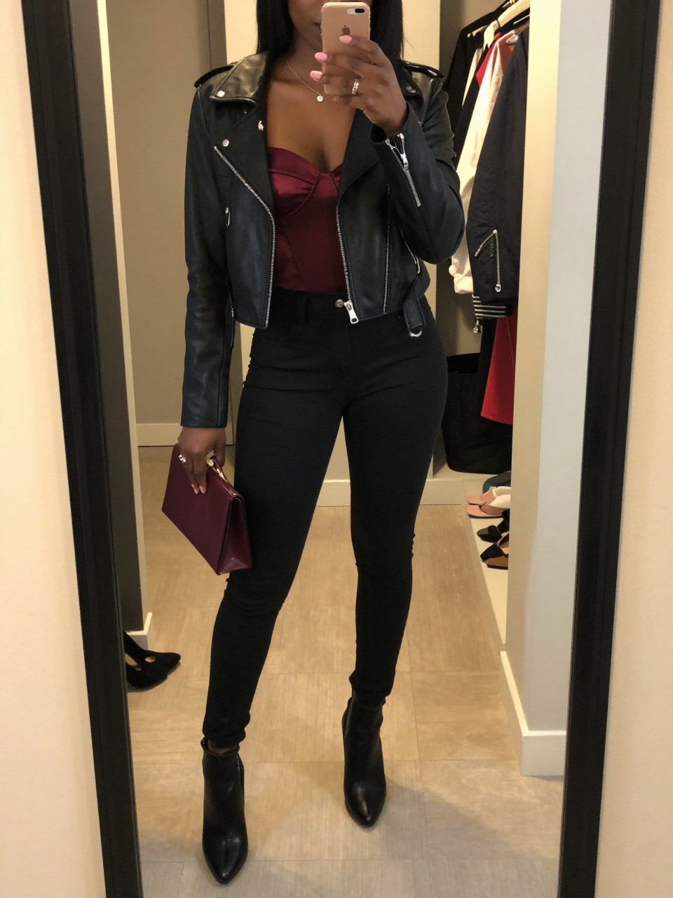 mirror selfie of a Black woman wearing the exact outfit described above, clearly showing a burgundy satin corset top, black coated skinny jeans, black cropped moto jacket, black heeled ankle boots, and burgundy mini clutch, holding a phone in front of a mirror, cropped at NECK so the face does not show, with a confident casual selfie pose, in a walk-in closet mirror, evening golden hour, including an open closet with clothes visible and a pair of heels on the floor nearby, latest iPhone photo quality.