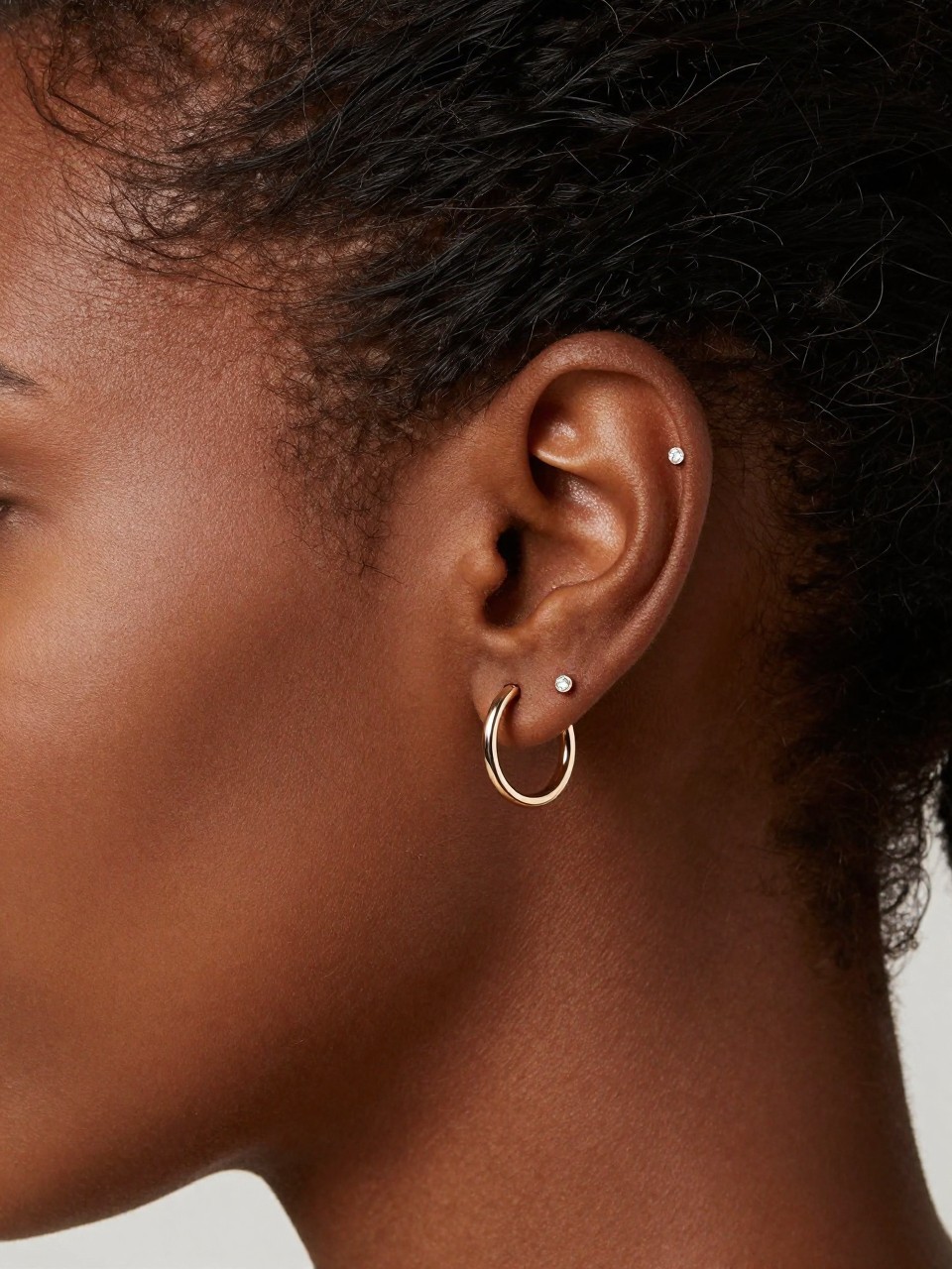 Close-up side portrait focused tightly on a Black woman’s ear with smooth espresso skin, hair pinned back, two inner conch piercings styled with a slim conch hoop and a tiny pavé conch stud, clean beauty editorial shot, soft light reflecting on the jewelry, highly realistic detail.