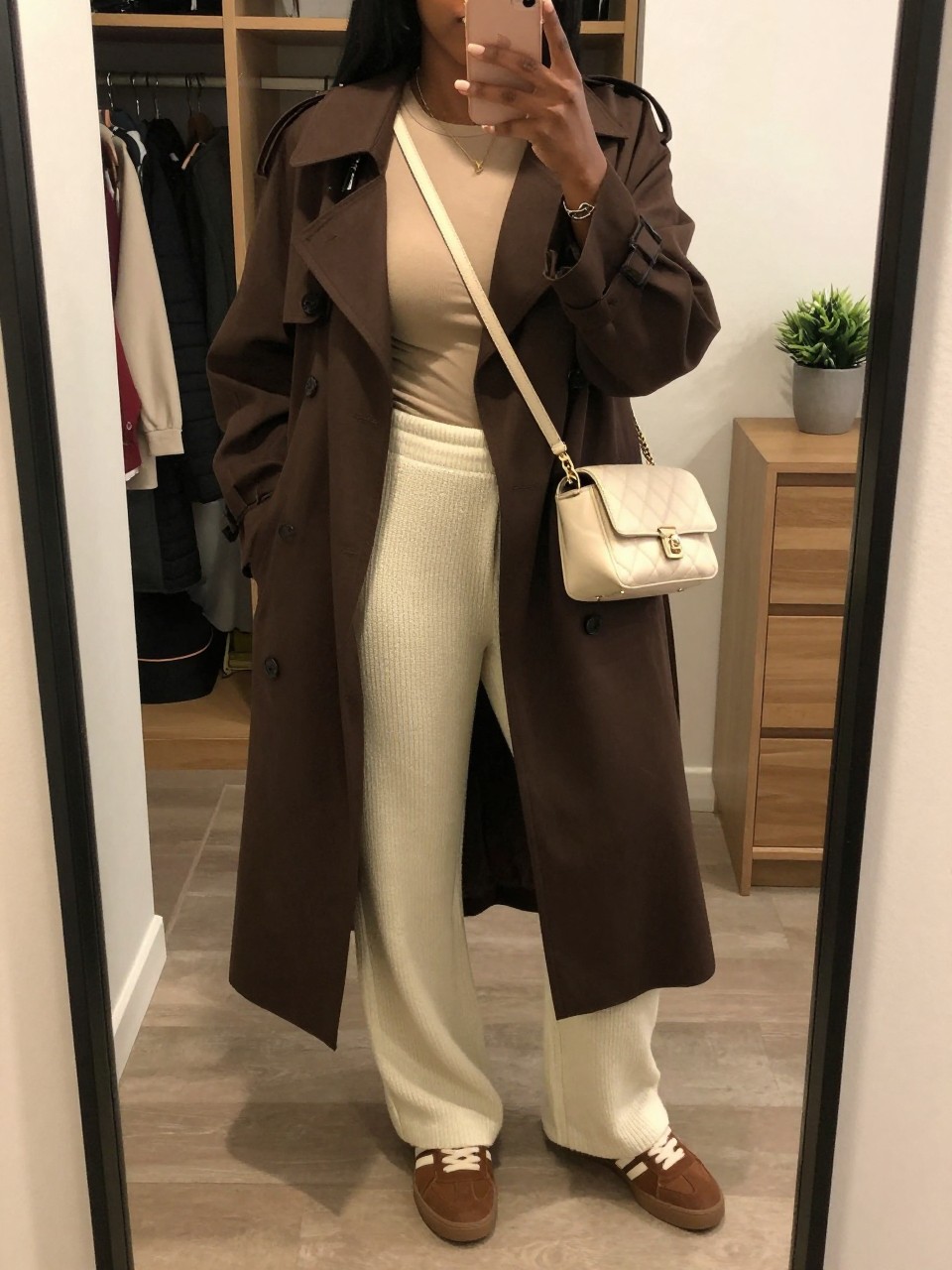 mirror selfie of a Black woman wearing the exact outfit described above, clearly showing a chocolate brown lightweight trench coat, beige fitted long-sleeve top, cream knit wide-leg pants, brown low-profile sneakers, and cream quilted crossbody bag, holding a phone in front of a mirror, cropped at NECK so the face does not show, with a relaxed casual selfie pose, in a walk-in closet mirror, warm moody lighting, including an open closet with clothes visible and a small plant on a dresser, latest iPhone photo quality.