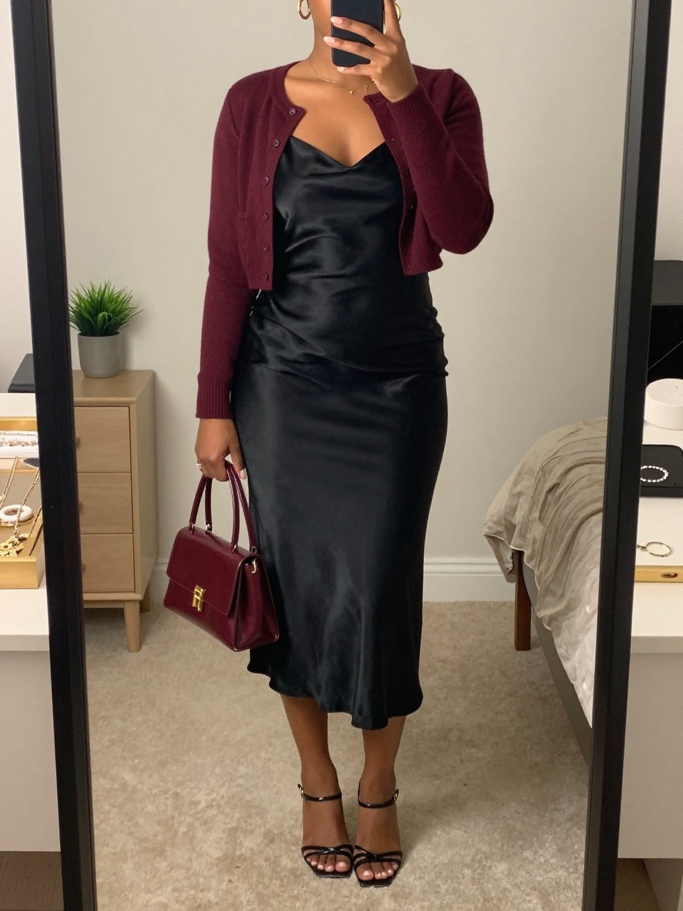 mirror selfie of a Black woman wearing the exact outfit described above, clearly showing a black satin slip midi dress, burgundy cropped button-up cardigan, black strappy kitten heels, burgundy mini handbag, and gold hoop earrings, holding a phone in front of a mirror, cropped at NECK so the face does not show, with a relaxed birthday selfie pose, in a feminine-themed bedroom, soft indoor lighting, including a small plant on a dresser and a jewelry tray on the vanity, latest iPhone photo quality.