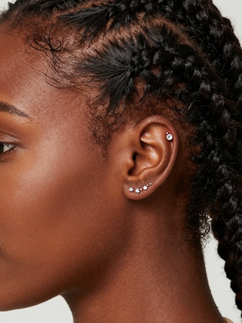 Editorial ear close-up of a Black woman with rich mahogany skin, sleek braids tucked behind the ear, one forward helix crystal stud and one tragus crystal stud forming a clean line near the face, soft luxury lighting, realistic beauty shot, crisp focus on front-of-ear piercings.