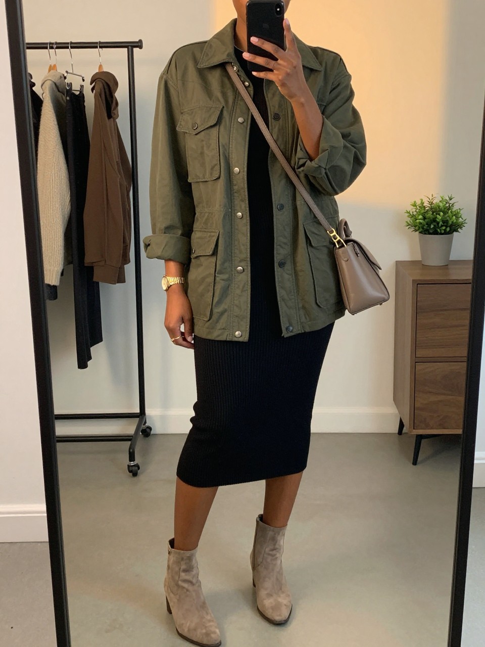mirror selfie of a Black woman wearing the exact outfit described above, clearly showing an olive green utility jacket with front pockets, black ribbed knit midi dress, beige suede ankle boots, taupe crossbody bag, and gold watch, holding a phone in front of a mirror, cropped at NECK so the face does not show, with a relaxed casual selfie pose, in a minimal dressing room, evening golden hour, including a clothing rack with garments behind and a small plant on a dresser, latest iPhone photo quality.