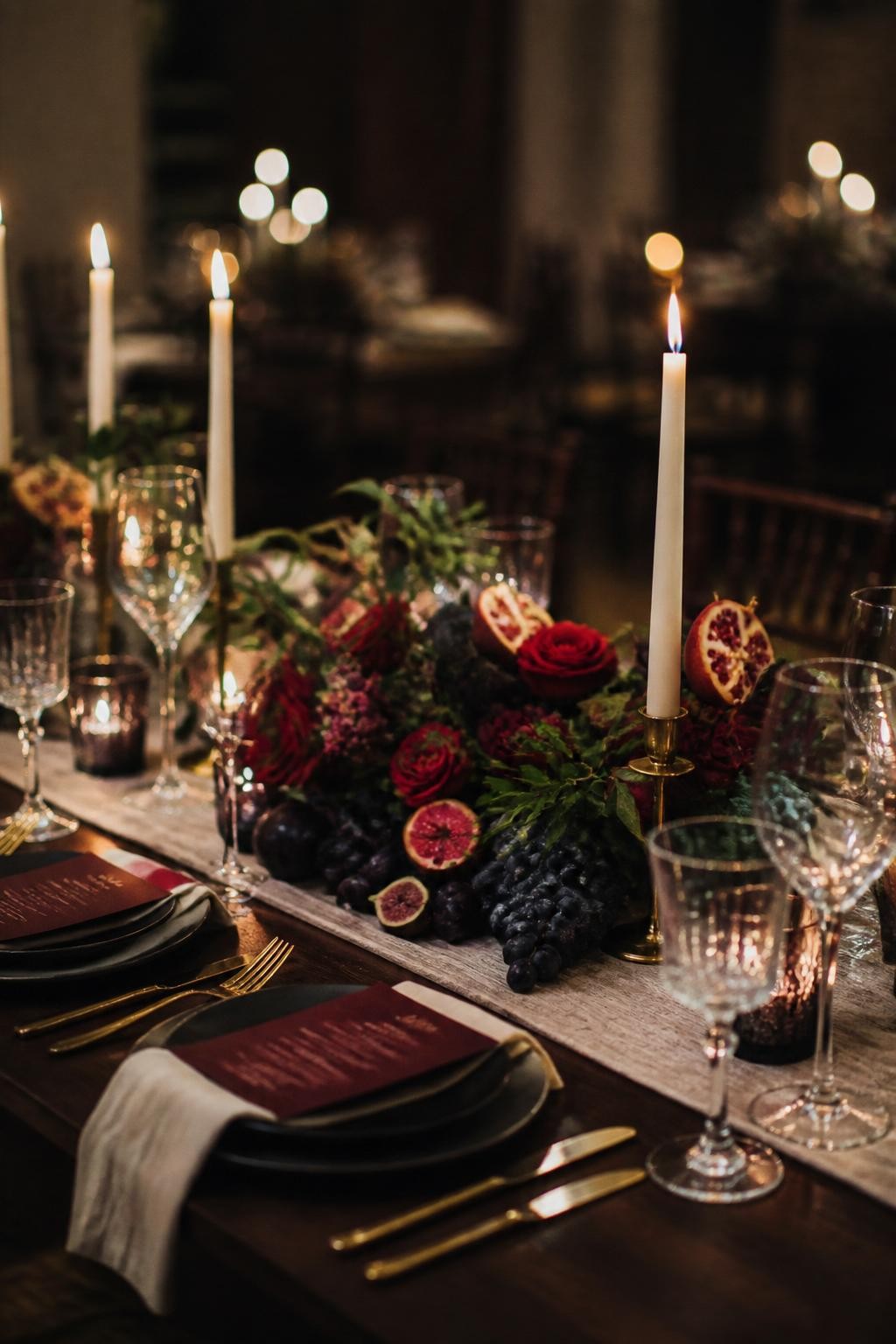 Realistic DSLR photo of a fall wedding tablescape with low centerpieces made of figs, black grapes, plums, pomegranates, ruby roses, amethyst scabiosa, emerald leaves, cream taper candles, gold flatware, linen runner, moody dinner reception.