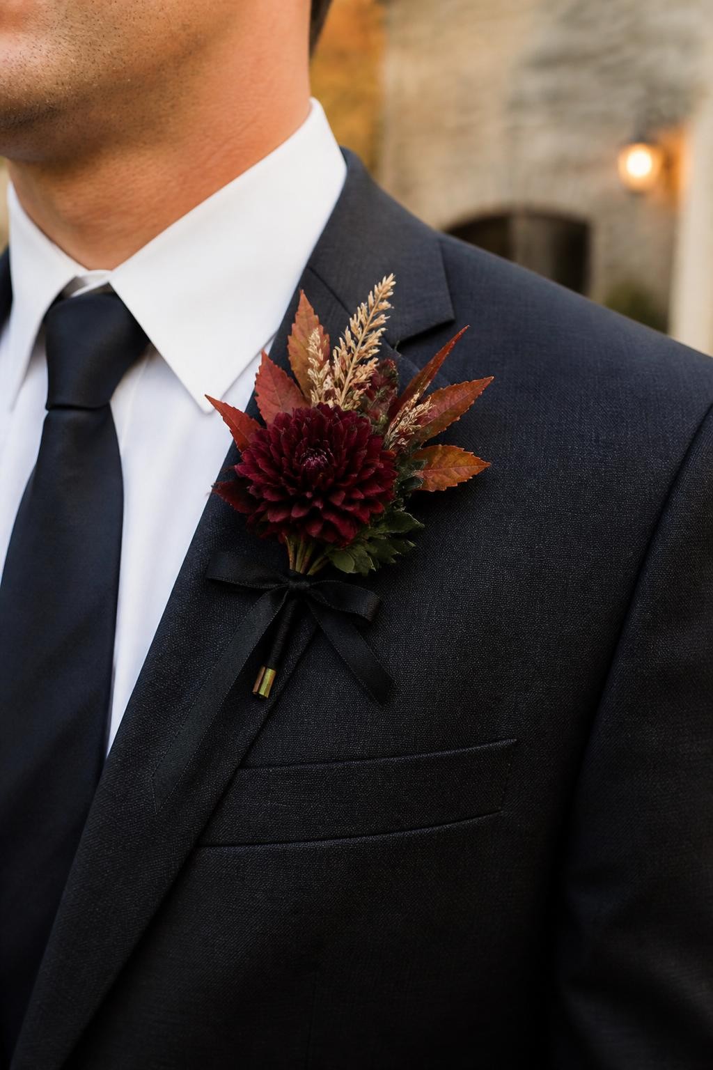 Realistic DSLR close-up of a groom’s charcoal suit lapel with a mini burgundy dahlia boutonniere, rust autumn leaves, tiny dried grass stems, dark green foliage, thin black silk ribbon, warm outdoor fall light, stone venue background softly blurred.