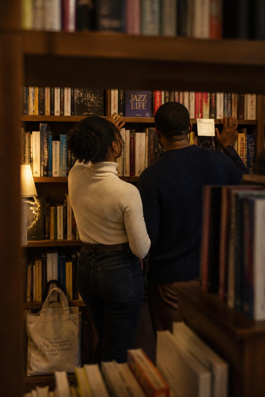 Image prompt: A warm bookstore couple photo taken from behind tall shelves, no faces shown, featuring an adult Black woman with dark brown skin wearing a fitted cream turtleneck, dark jeans, and gold bangles, standing beside her partner in a navy sweater, both facing a wall of books with their backs to the camera, shoulders touching, her hand reaching for a book while his hand rests lightly on the shelf near hers, faces completely turned away and hidden, warm reading lamps, wooden shelves, canvas tote on the floor, realistic paper texture, quiet faceless date photography, DSLR lifestyle style.