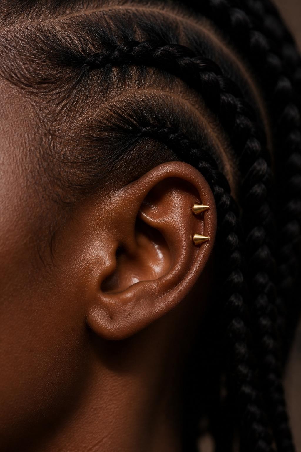 Extreme close-up of a Black woman’s ear with warm dark skin, cornrows pulled back cleanly, two mid-helix piercings stacked closely with tiny polished gold spike studs, soft moody editorial lighting, realistic skin texture, crisp detail on cartilage piercings, minimal background.