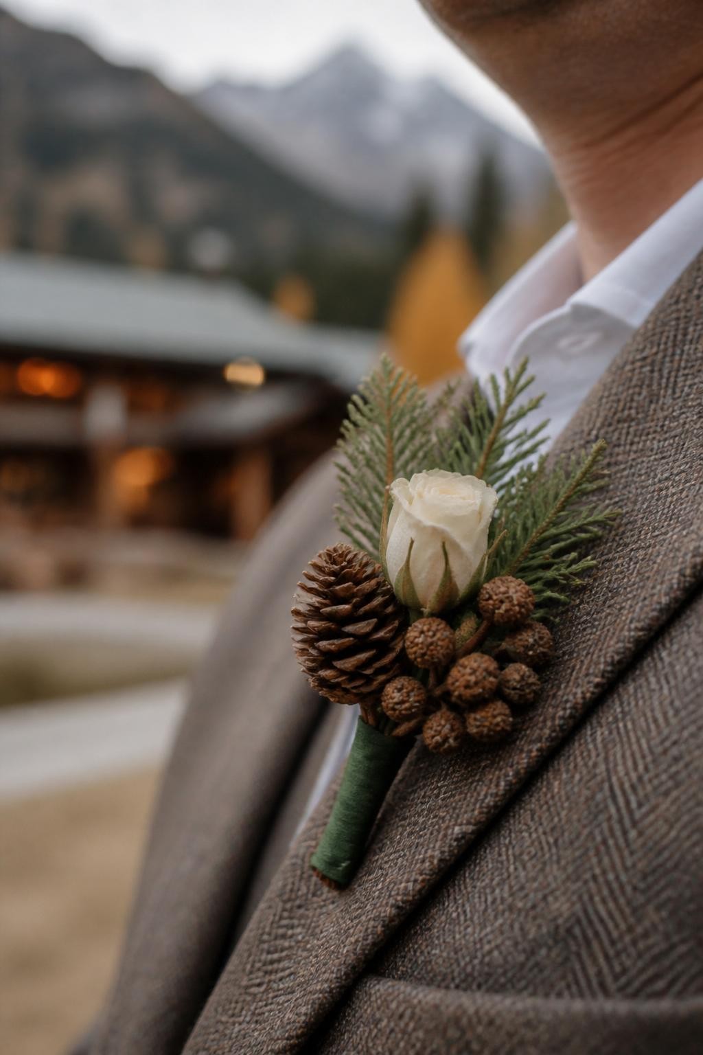Realistic DSLR macro photo of a fall mountain wedding boutonniere with a tiny pinecone, cedar sprig, cream rosebud, brown seed pods, forest green ribbon wrap, pinned to a tweed suit lapel, mountain lodge background softly blurred, cozy autumn styling.