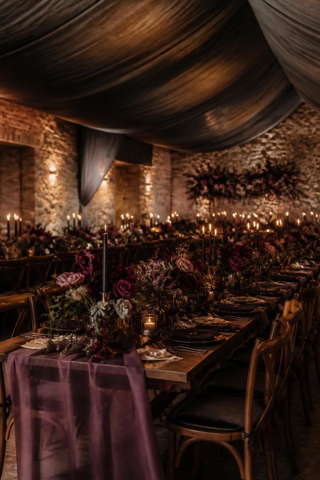 Realistic DSLR photo of a fall wedding reception room with smoky gray ceiling draping, long candlelit tables, burgundy and plum floral centerpieces, black taper candles, walnut chairs, warm amber lighting, stone walls, soft shadows, no visible faces, dramatic romantic dinner setting.