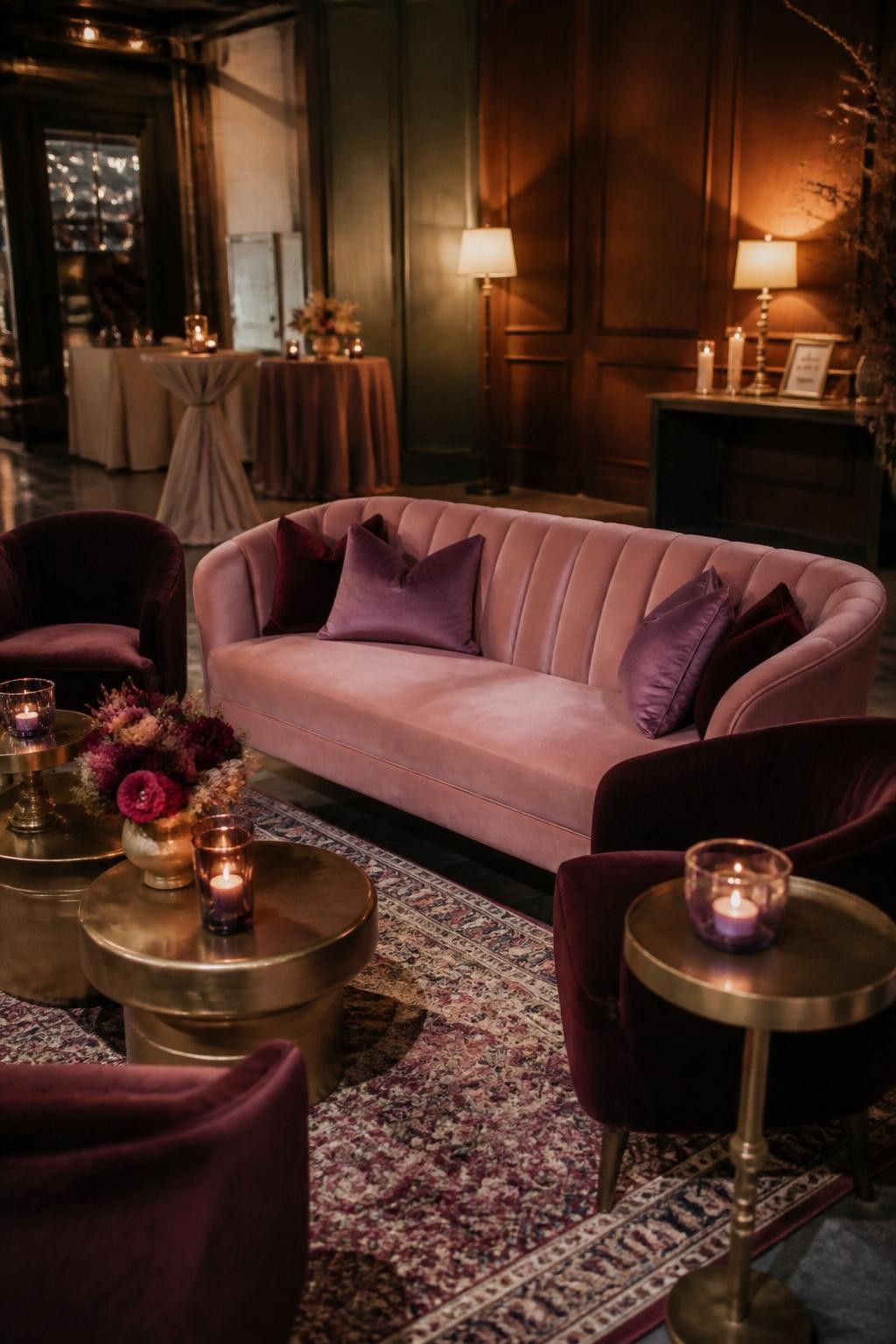 Realistic DSLR photo of a fall wedding lounge area with dusty pink velvet sofa, plum velvet chairs, amethyst glass candle holders, brass cocktail tables, patterned rug, mauve and burgundy flower arrangement, warm table lamps, cozy indoor reception corner, no visible faces.