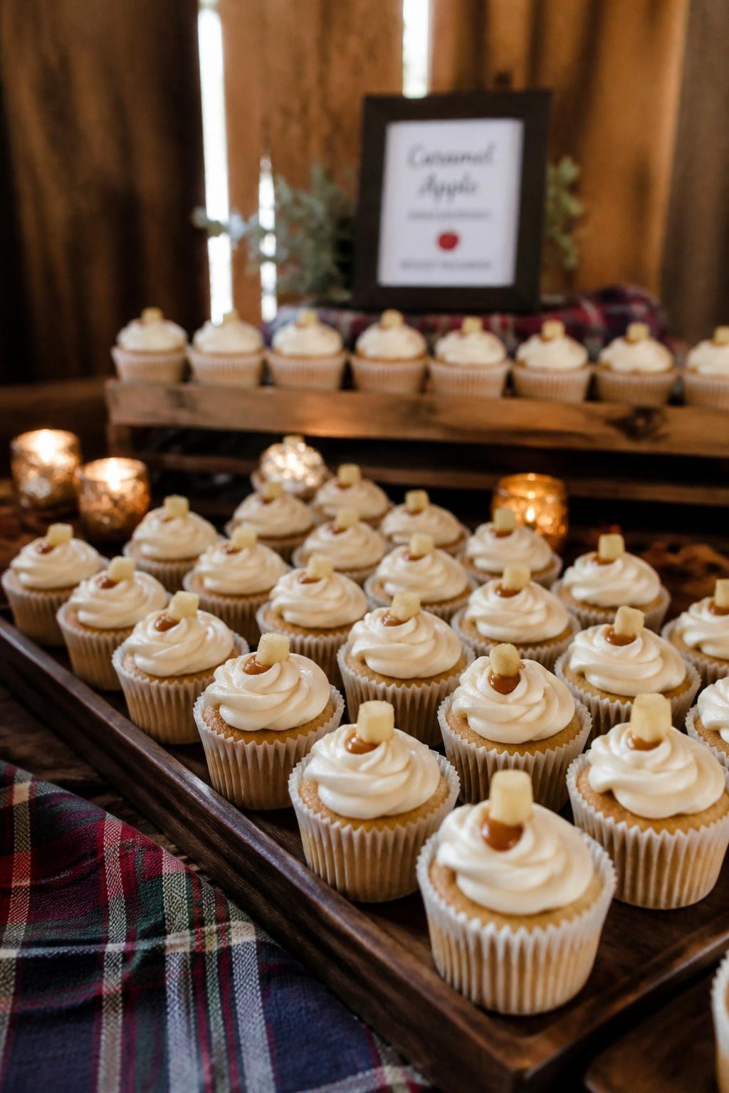 Realistic DSLR photo of mini fall wedding cupcakes in neat rows, caramel apple flavor, ivory frosting swirls, tiny caramel dots, small apple cube garnish, wooden trays, plaid napkin detail, amber candles, barn wedding dessert table.