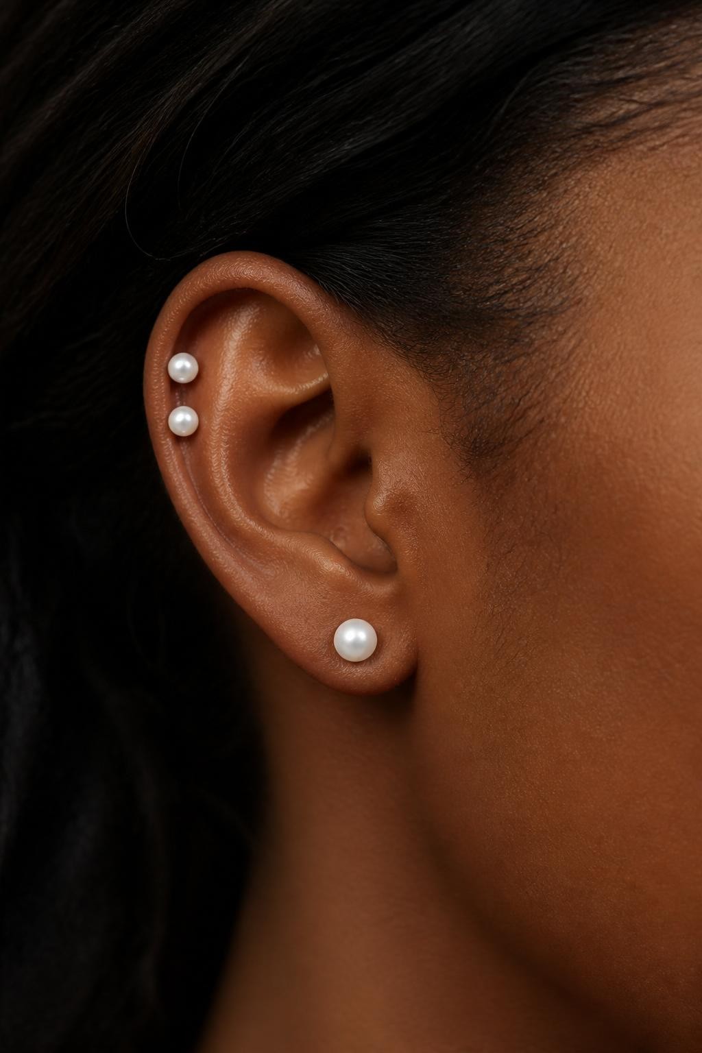 Close-up side view of an ear with deep brown skin tone, triple piercing arrangement featuring two small pearl studs on the upper ear and one pearl lobe stud, hair tucked back, soft natural light, realistic skin texture, elegant editorial beauty photography, full focus on the pearls and ear.