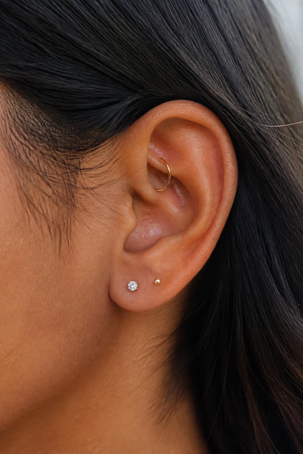 Side close-up of a small ear with warm brown skin tone, one delicate gold hoop through the rook and two tiny lobe studs below, hair tucked behind the ear, soft daylight, realistic editorial photo, clear focus on the inner ear hoop and neat lobe piercings.