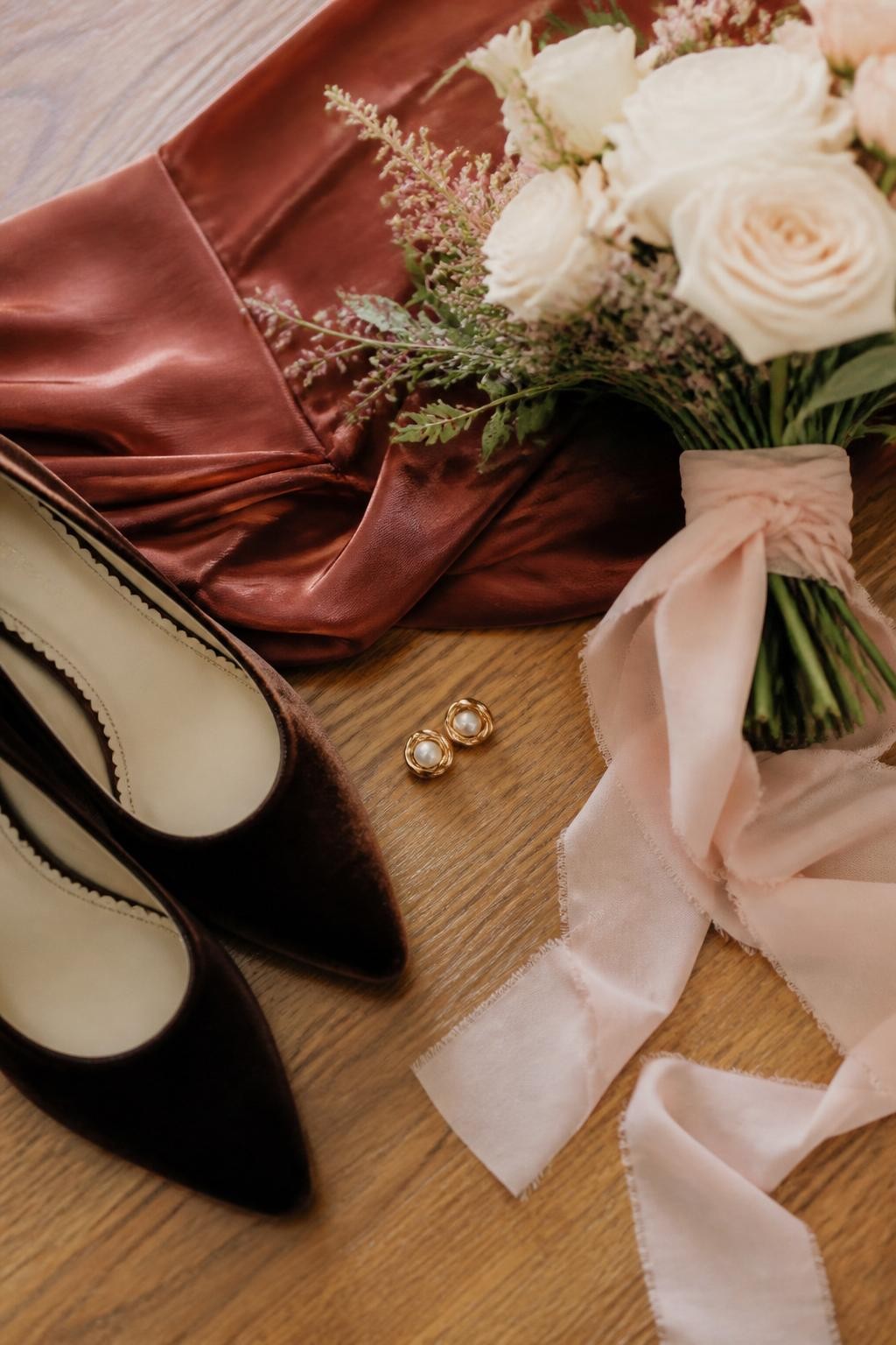 Realistic DSLR photo of a fall bridal party detail scene, rust bridesmaid dress fabric, ballet pink bouquet ribbon, espresso velvet shoes, warm ivory roses, small gold earrings, wood floor background, soft morning light, elegant wedding styling flat lay.