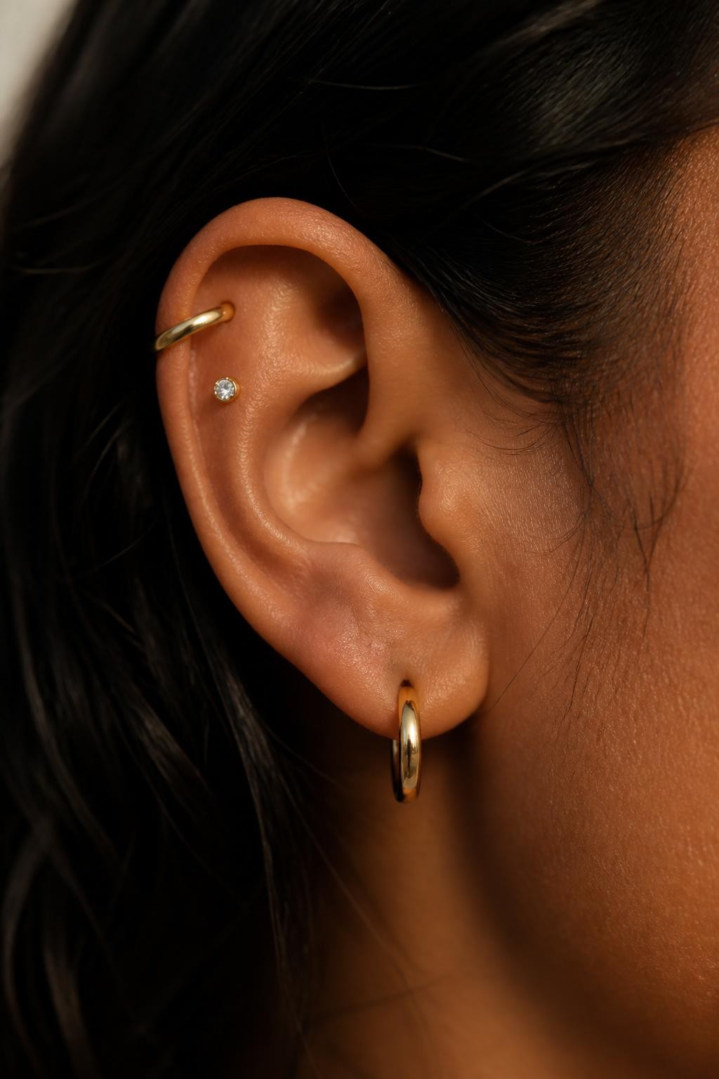 Close-up side view of a small ear with warm brown skin tone, upper helix decorated with one slim gold ear cuff and one tiny crystal helix stud, lobe styled with a petite gold huggie, hair tucked behind ear, soft warm natural light, realistic DSLR beauty photography, clear focus on the upper ear crown effect.