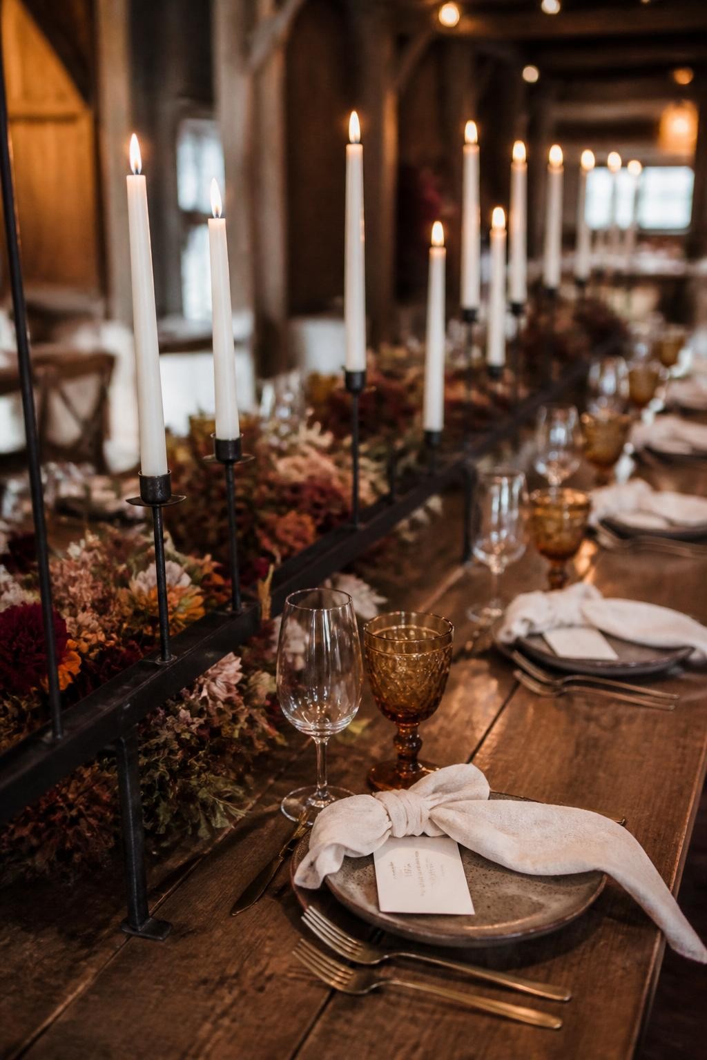 Realistic DSLR photo of a rustic fall wedding table with a long black iron candle rail running down the center, cream taper candles, low rust and burgundy flowers, walnut wood table, stone plates, linen napkins, amber glassware, moody barn reception lighting.