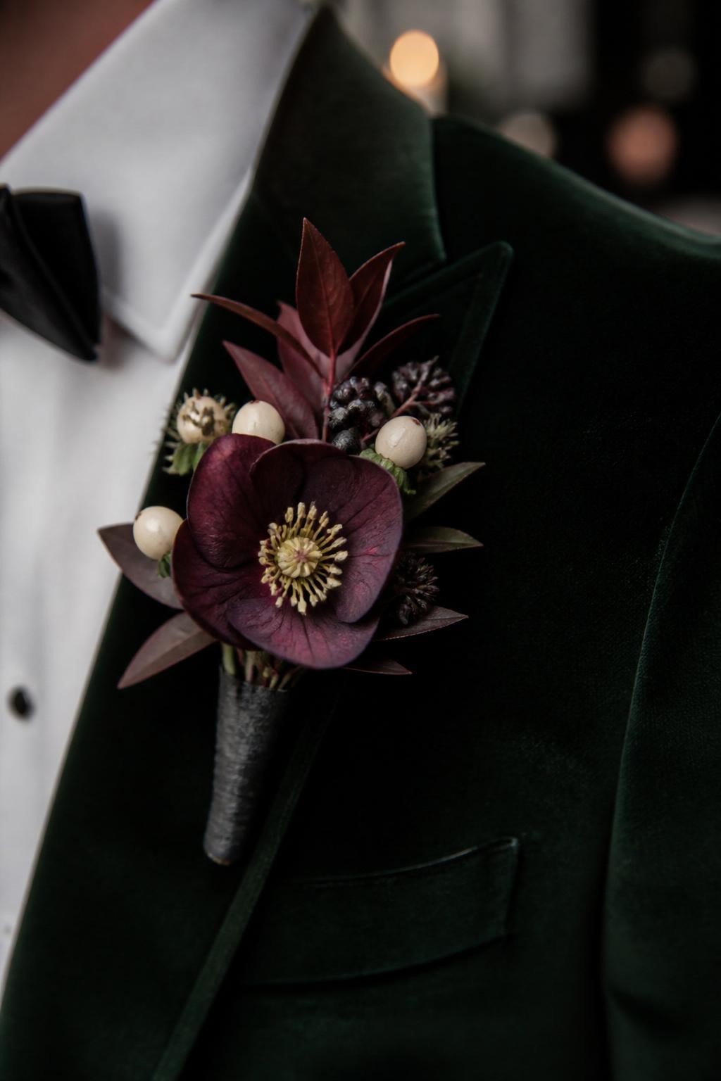 Realistic DSLR macro photo of a fall wedding boutonniere with plum hellebore, tiny cream berries, dark burgundy foliage, black silk ribbon wrap, pinned to a deep green velvet suit lapel, moody indoor candlelit wedding setting, groom face cropped out.