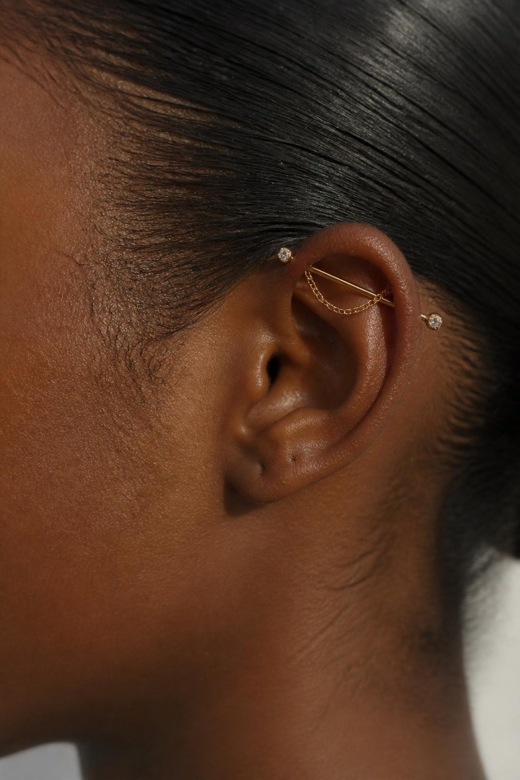 Side portrait of an ear with deep brown skin tone, upper ear styled with a delicate industrial bar and a very fine gold chain draping lightly along the helix, tiny crystal ends, hair slicked back, realistic natural light, editorial close-up, clean focus on the top ear jewelry.