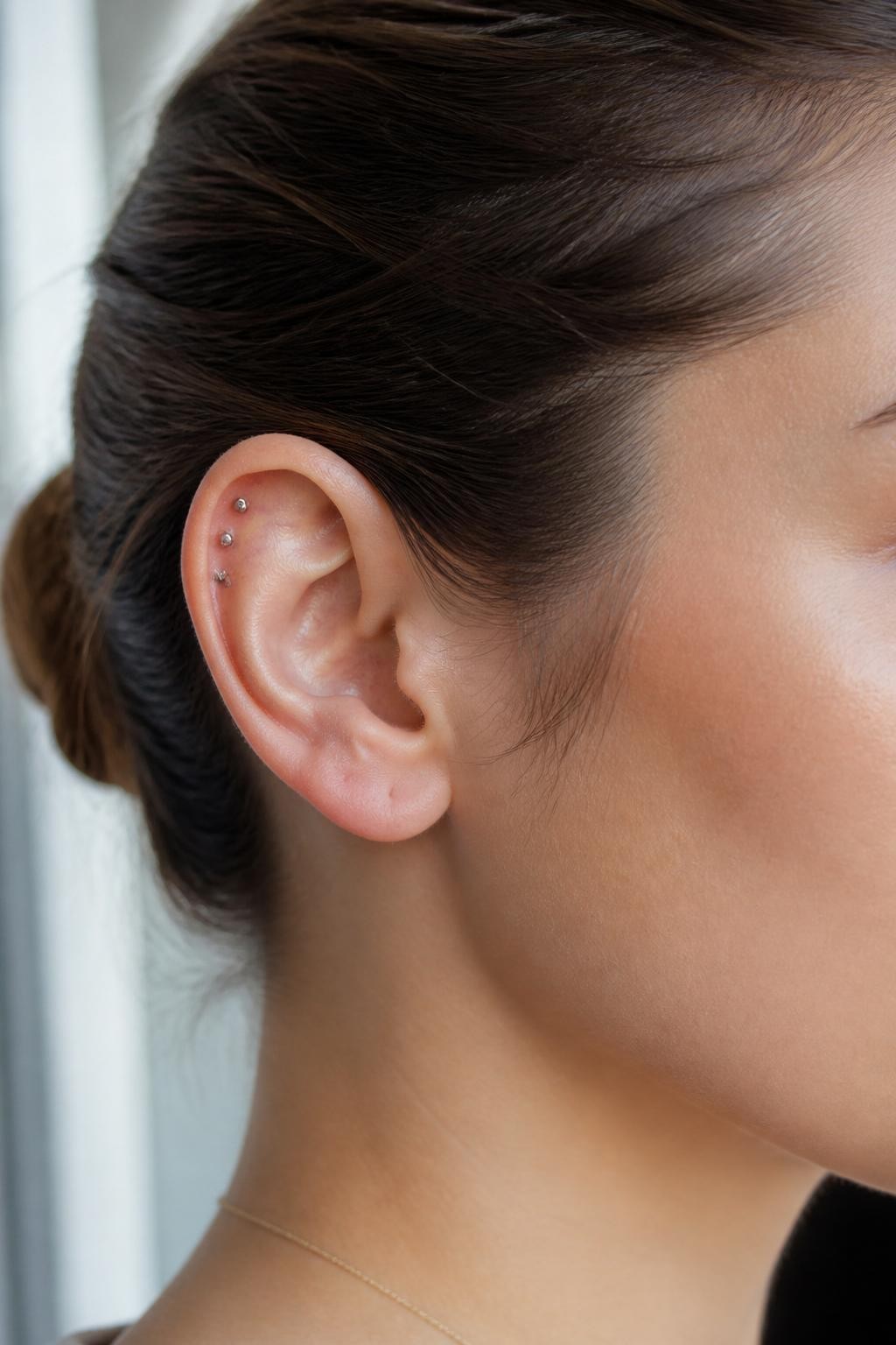 Side portrait close-up of an ear with neutral fair skin tone, three very small silver studs lined vertically on the forward helix near the front of the ear, sleek hair tucked back, soft window light, realistic beauty editorial style, sharp focus on the front upper ear.
