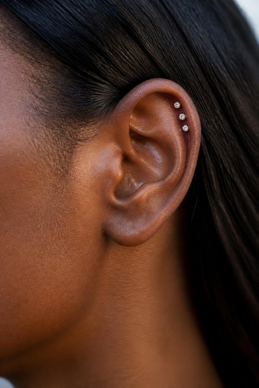 Side portrait close-up of a small ear with deep brown skin tone, hair tucked neatly behind the ear, three tiny crystal studs placed closely along the upper helix in a soft rising line, petite gold jewelry, natural daylight, realistic skin texture, clean editorial beauty photography, sharp focus on the ear shape and piercings.