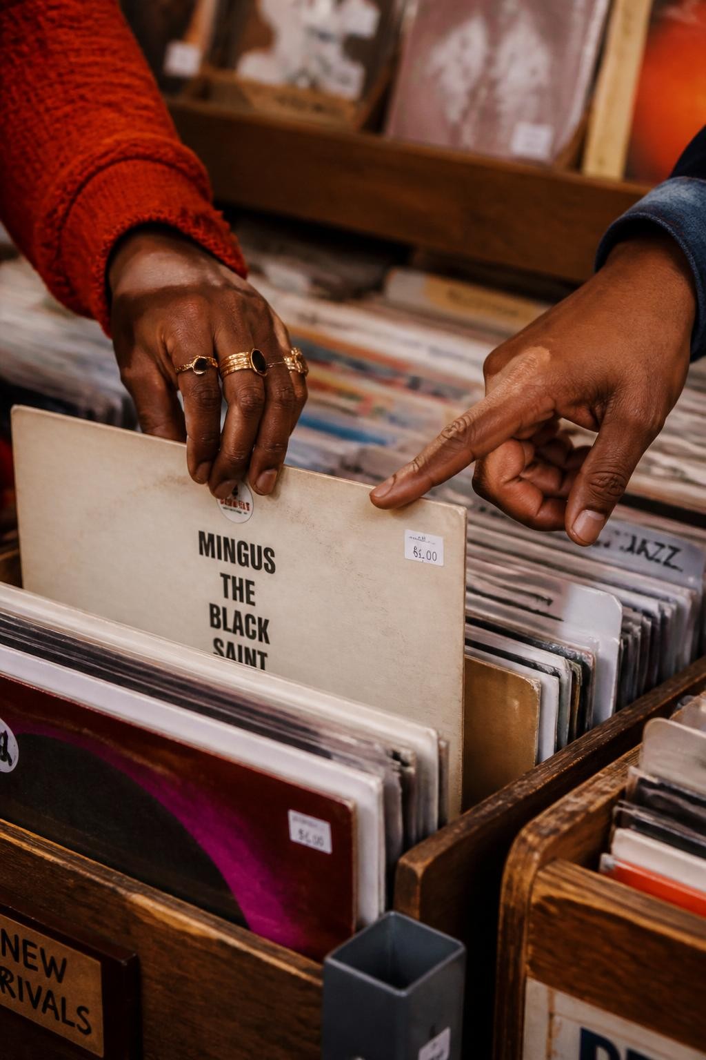 Image prompt: A creative DSLR close-up of an adult couple’s hands flipping through vinyl records inside a small record shop, no faces or upper bodies shown, featuring a Black woman with deep brown skin wearing stacked gold rings and a red knit sleeve, her hand pulling out a record sleeve while her partner’s hand points to the album cover, wooden record bins, colorful album covers, small price stickers, warm shop lighting, realistic paper edges, slight dust on the records, cozy music-date mood, faceless couple lifestyle photography.