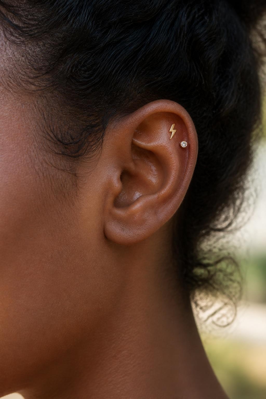 Side portrait close-up of a small ear with deep brown skin tone, flat upper cartilage styled with a tiny gold lightning bolt stud and one small crystal accent nearby, hair pulled back, soft warm daylight, realistic skin texture, editorial DSLR photography, sharp focus on the upper ear jewelry.