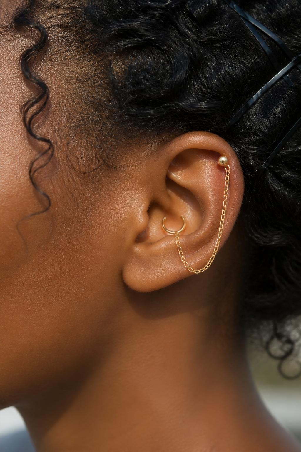 Close-up side portrait of a Black woman’s ear with deep golden-brown skin, natural curls pinned back, one inner conch hoop and one upper helix stud connected by a fine gold chain, editorial close-up, soft daylight, realistic jewelry shine, sharp focus on the ear styling and chain detail.