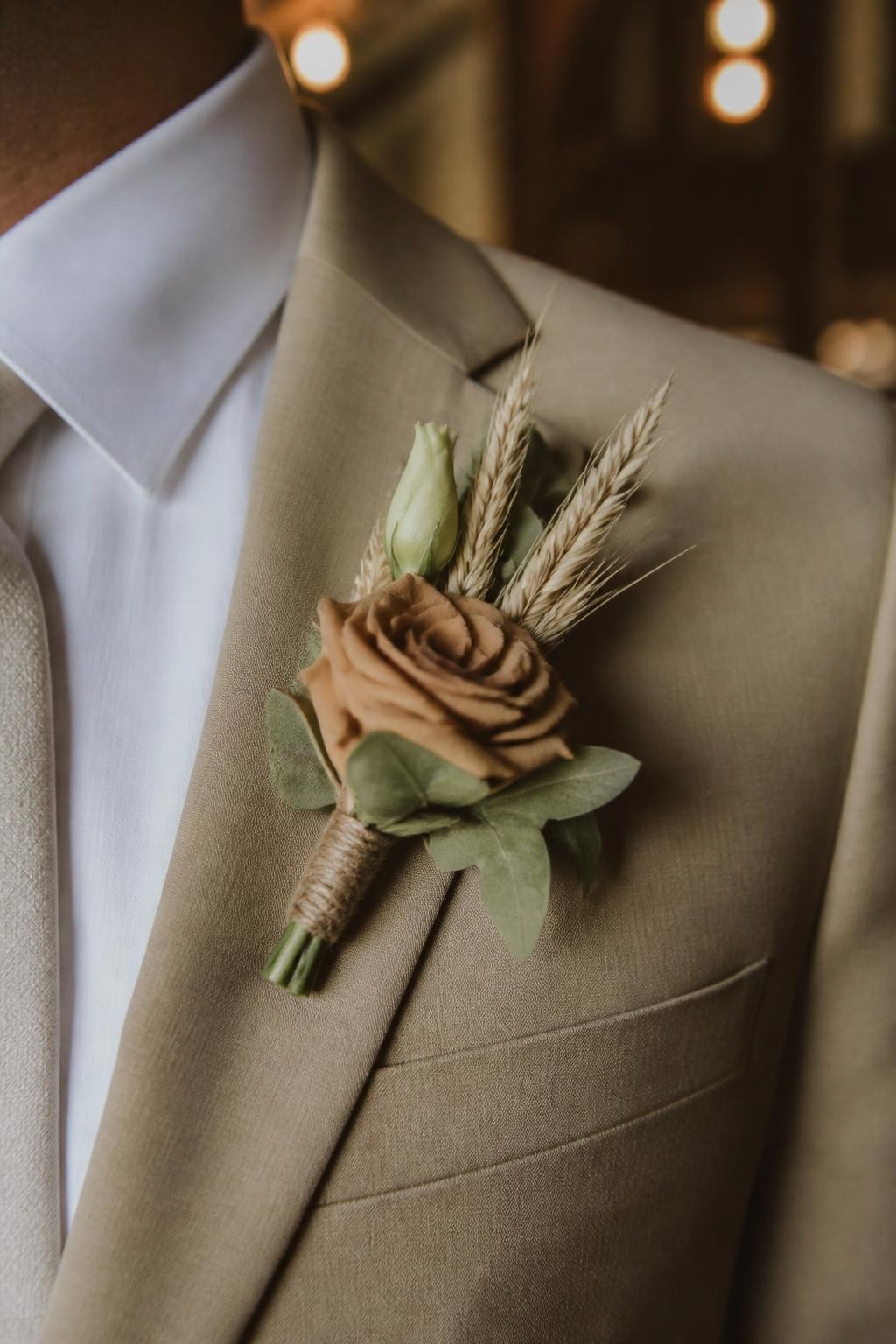 Realistic DSLR photo of a groom’s tan suit lapel with a toffee rose boutonniere, dried wheat, cream lisianthus bud, soft sage leaves, natural twine wrap, warm barn wedding light, rustic fall wedding styling, face not visible.