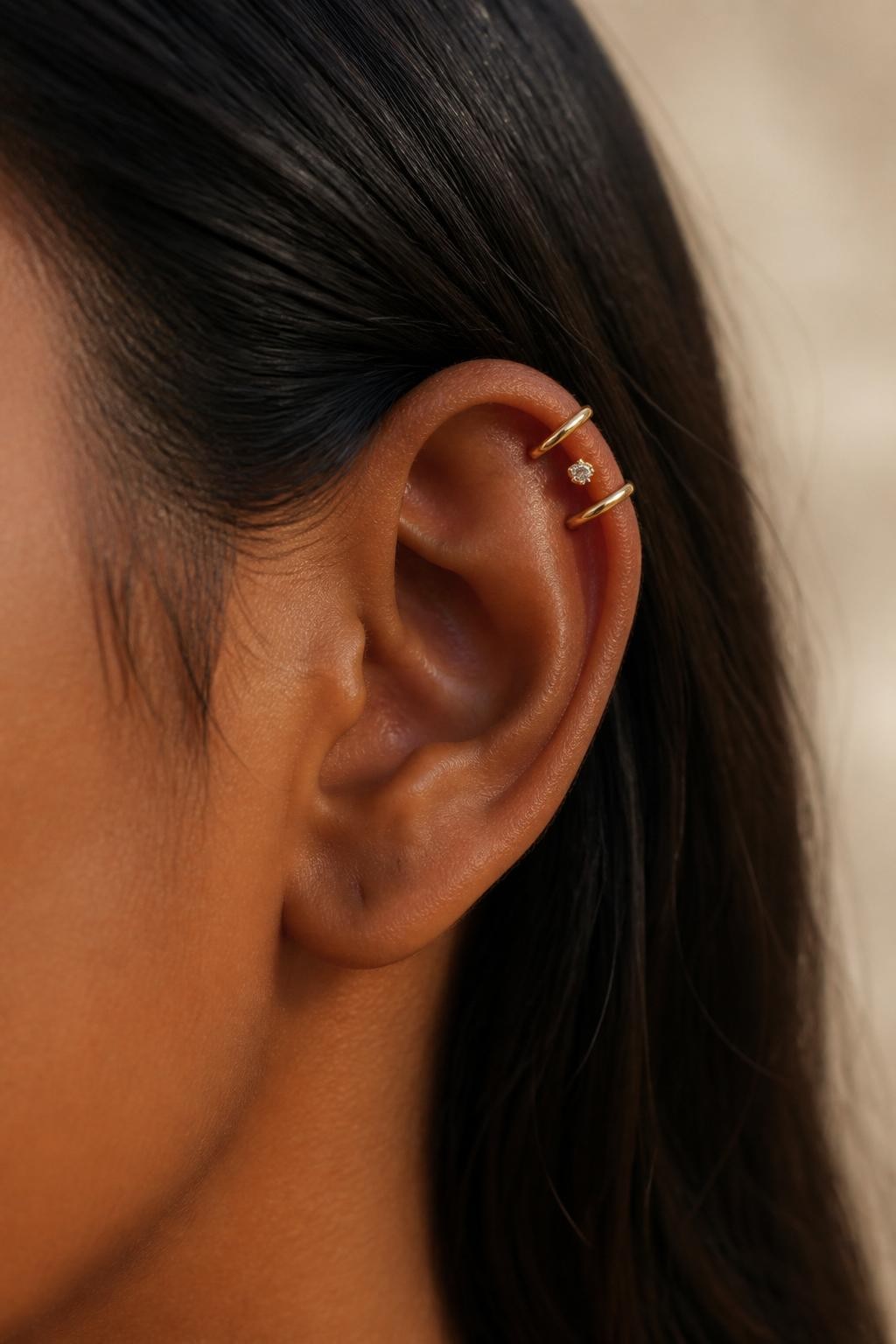 Side close-up of an ear with warm dark brown skin tone, upper helix styled with two slim gold cuffs and one tiny pierced stud between them, crown-like placement along the top edge of the ear, hair tucked behind ear, soft beige background, natural daylight, realistic editorial photo, focus on upper cartilage.