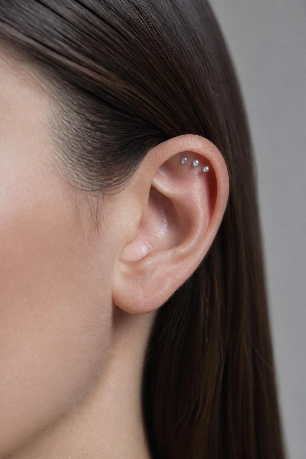 Close-up side view of an ear with light neutral skin tone, flat upper cartilage styled with three tiny silver studs in a straight horizontal rail, sleek tucked hair, cool natural daylight, simple grey background, realistic editorial beauty photography, sharp detail on the upper ear.