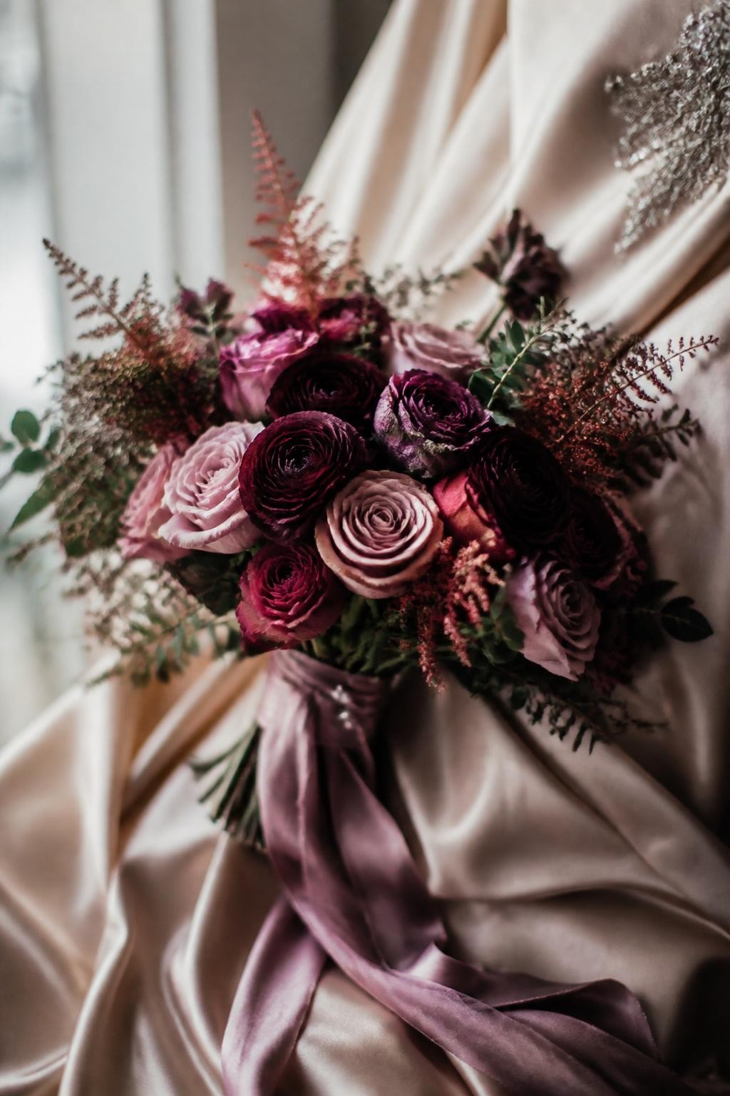 Realistic DSLR close-up of a fall bridal bouquet with mulberry ranunculus, dusty rose garden roses, smoked mauve ribbon, champagne silk dress fabric in the background, small pearl pins, soft window light, romantic editorial wedding photography, elegant but natural.