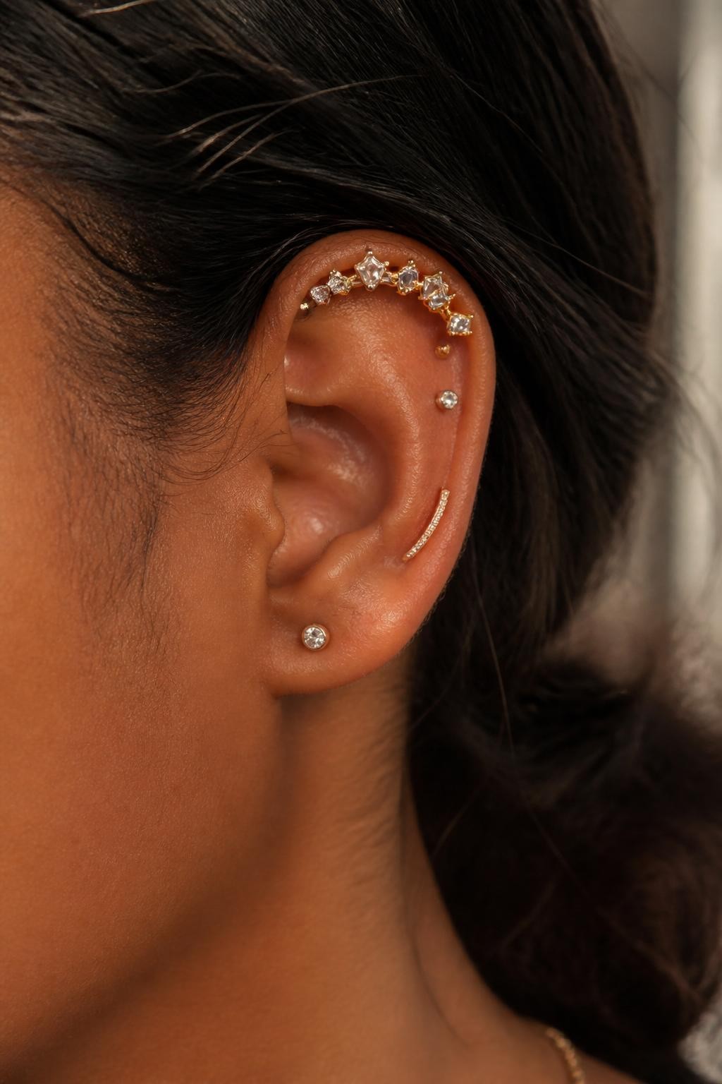 Side portrait close-up of an ear with warm brown skin tone, upper helix and flat area styled with a mixed metal crown arc, tiny gold stud, tiny silver stud, rose-gold curved bar, and small crystal accent arranged along the upper ear, hair tucked back, soft warm daylight, realistic DSLR editorial style.