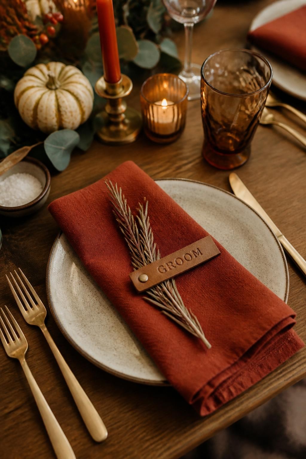 Realistic DSLR close-up of a rustic fall wedding place setting with handmade stoneware plate, folded rust linen napkin, small leather name tag, brass flatware, dried rosemary sprig, wooden table, amber glass, soft candlelight, detailed editorial wedding photography.