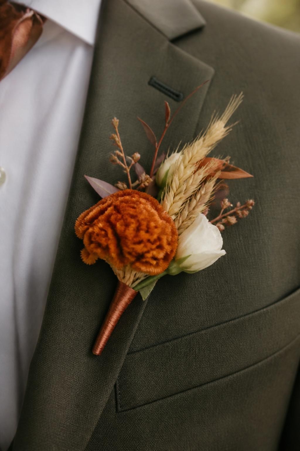 Realistic DSLR macro shot of a fall wedding boutonniere pinned to an olive green suit lapel, rust celosia, cream lisianthus bud, dried wheat, tiny brown leaves, copper silk ribbon wrap, warm outdoor ceremony light, no face visible.