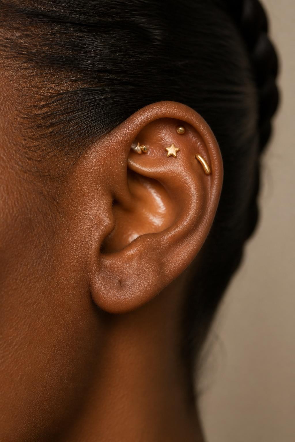 Close-up side view of an ear with rich dark brown skin tone, upper flat area styled with tiny sculpted gold studs in uneven constellation placement, one small star stud, one tiny dot stud, one curved gold piece, hair pulled back, warm natural light, realistic DSLR photo, clean background, sharp focus on upper ear jewelry.
