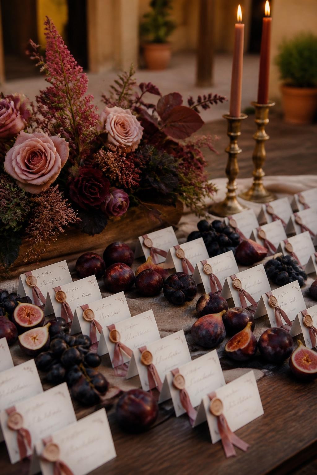 Realistic DSLR photo of a fall wedding escort card display with figs, black grapes, plums, and small cream name cards tied with dusty pink ribbon, low mauve floral arrangements, brass candlesticks, dark wood table, linen runner, warm courtyard light.