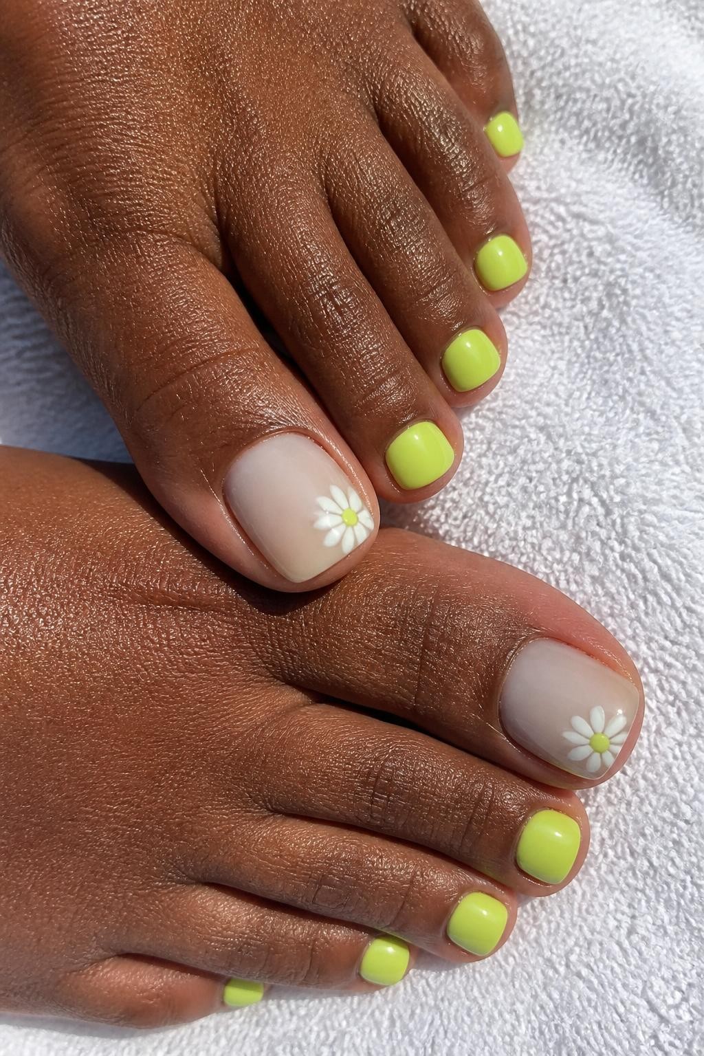 Close-up realistic photo of a pedicure on deep brown skin, glossy lime sorbet polish on smaller toes, milky nude big toenails with tiny white daisy nail art and lime centers, feet resting on a white towel, bright natural daylight, no face visible.