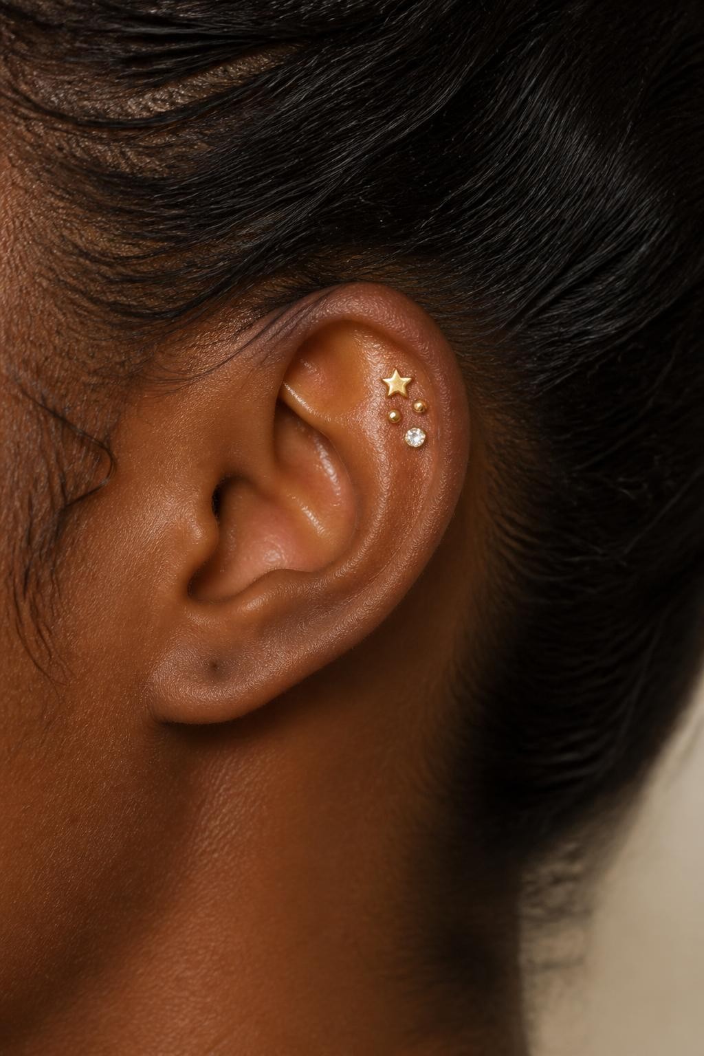 Close-up side view of a small ear with rich dark brown skin tone, upper flat cartilage styled with a tiny star stud, one small dot stud, and one mini crystal in a compact cluster, hair pulled back softly, warm natural light, realistic DSLR beauty shot, simple background, focus on the flat piercing arrangement.