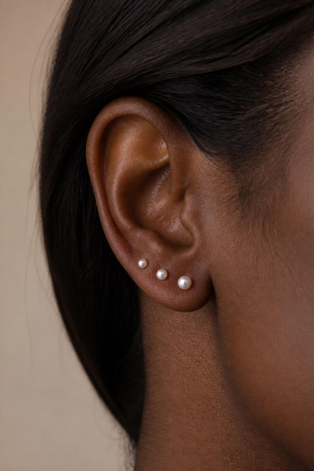 Side close-up of a small ear with rich dark brown skin tone, three tiny pearl studs placed along the lobe in a soft curved line, hair tucked behind the ear, soft beige background, natural light, realistic editorial beauty photography, sharp focus on the pearl piercings.