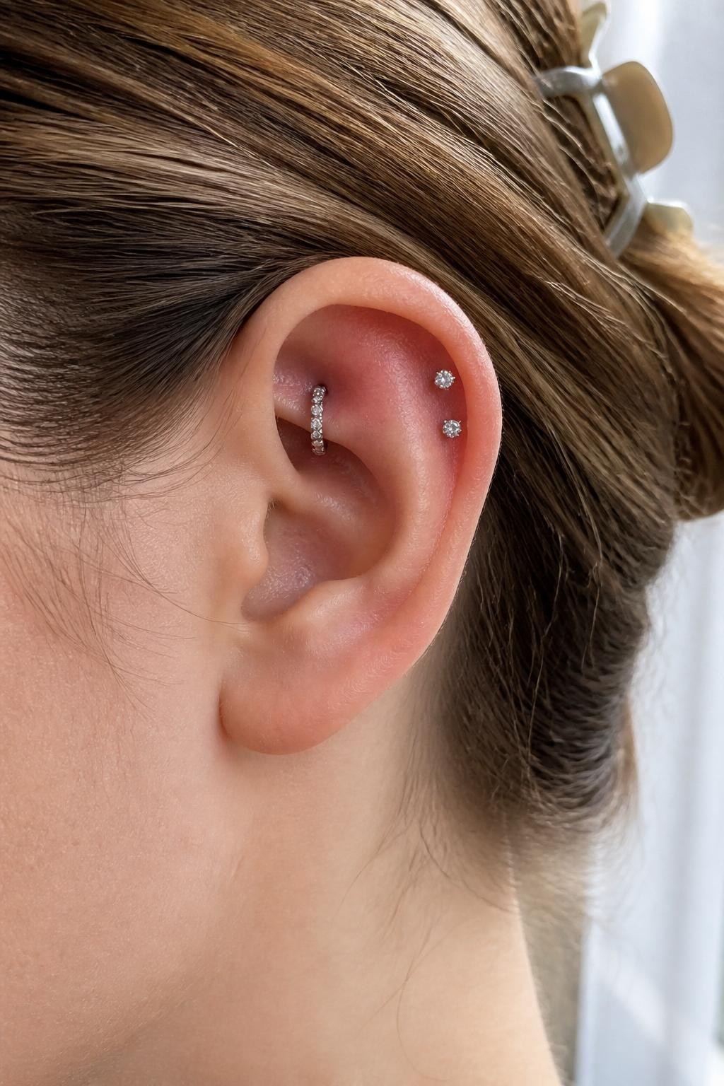 Side close-up of an ear with fair neutral skin tone, small silver gem hoop in the rook, two tiny crystal studs placed on the flat upper cartilage, hair tucked back with a simple clip, soft natural window light, realistic editorial beauty shot, clean focus on inner and upper ear jewelry.
