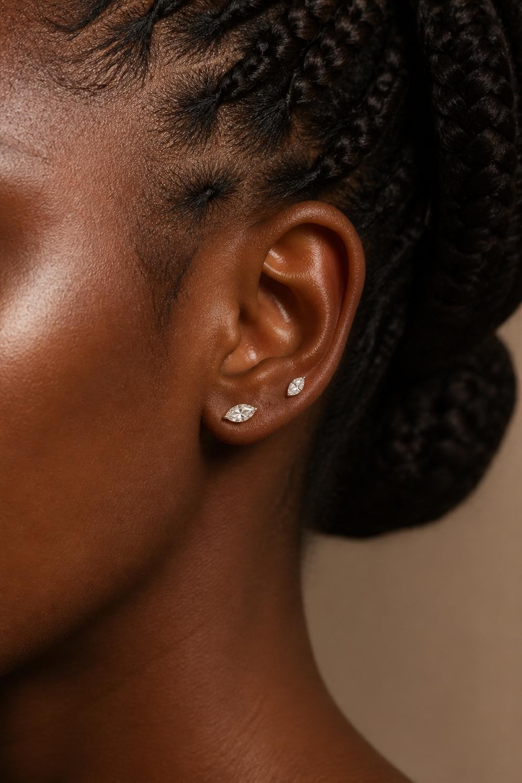 Beauty close-up of a Black woman’s side profile focused on the ear, smooth dark-brown skin, braids pulled into a bun, one flat piercing with a marquise crystal stud and one high lobe piercing with a matching smaller marquise stud, soft luxe lighting, realistic beauty editorial style, ear centered in frame.