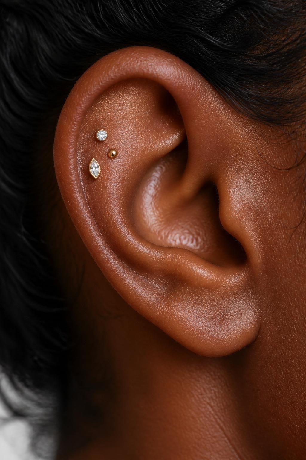 Close-up side view of an ear with rich dark brown skin tone, three tiny studs arranged in a soft triangle on the flat upper cartilage, one crystal, one gold bead, one marquise-shaped stud, hair pulled back, natural light, realistic DSLR beauty shot, full focus on the flat piercing arrangement.