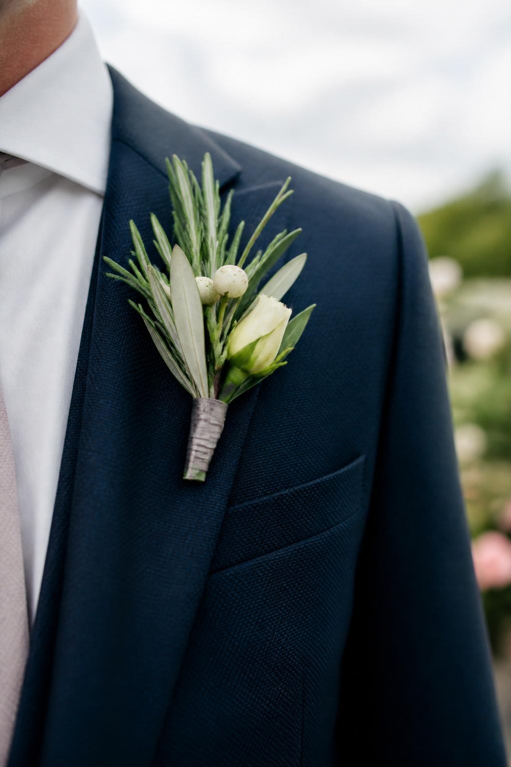 Realistic DSLR close-up of a fall wedding boutonniere made with rosemary sprigs, olive leaves, small cream berries, tiny ivory rosebud, wrapped in taupe silk ribbon, pinned to a navy suit lapel, outdoor garden wedding background, soft cloudy daylight.
