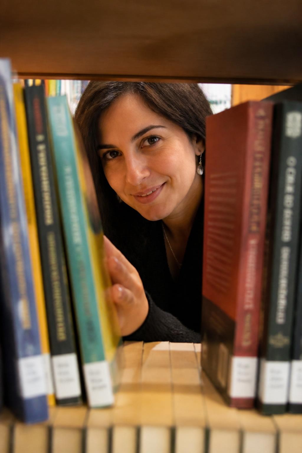 Image prompt: A playful DSLR library portrait of an adult woman with olive skin seen through a narrow gap between two rows of books on a shelf, wearing a soft black cardigan and delicate earrings, her face partly framed by book spines as she smiles gently through the opening, one hand visible pushing a book slightly aside, warm shelf light, colorful book edges close to the camera, background softly blurred, realistic paper texture, cute hidden library photo, human candid photography with a creative shelf-framed composition.
