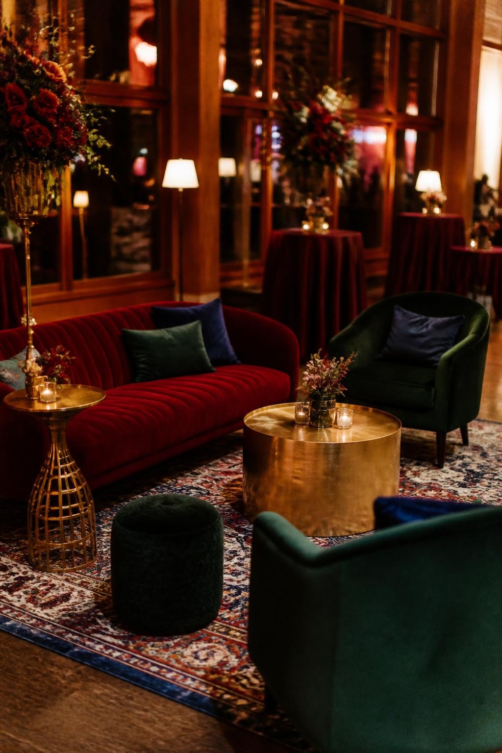 Realistic DSLR photo of a fall wedding lounge area with garnet red velvet sofa, emerald green armchairs, sapphire pillows, brass cocktail tables, patterned vintage rug, low lamps, dark floral arrangement, warm indoor reception lighting, cozy jewel tone wedding lounge.