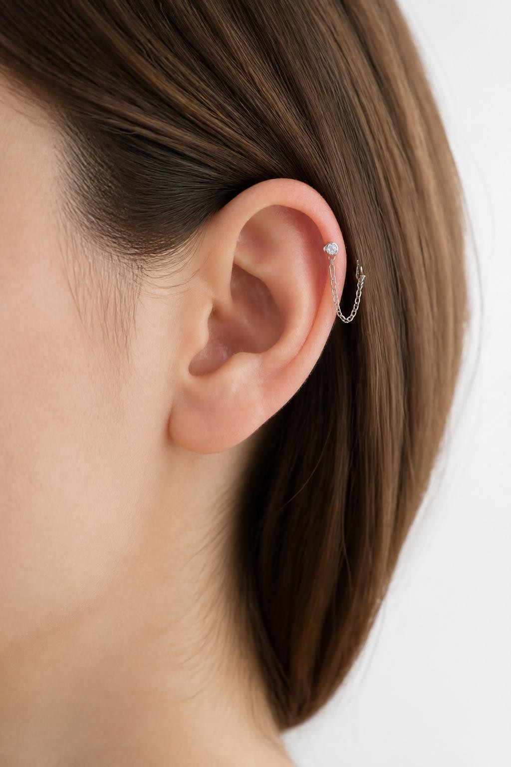 Side view close-up of an ear with light neutral skin tone, double upper helix piercing connected with a thin silver mini chain, two tiny crystal studs holding the chain, hair softly tucked behind ear, clean white background, natural light, realistic beauty photography, clear focus on upper helix.
