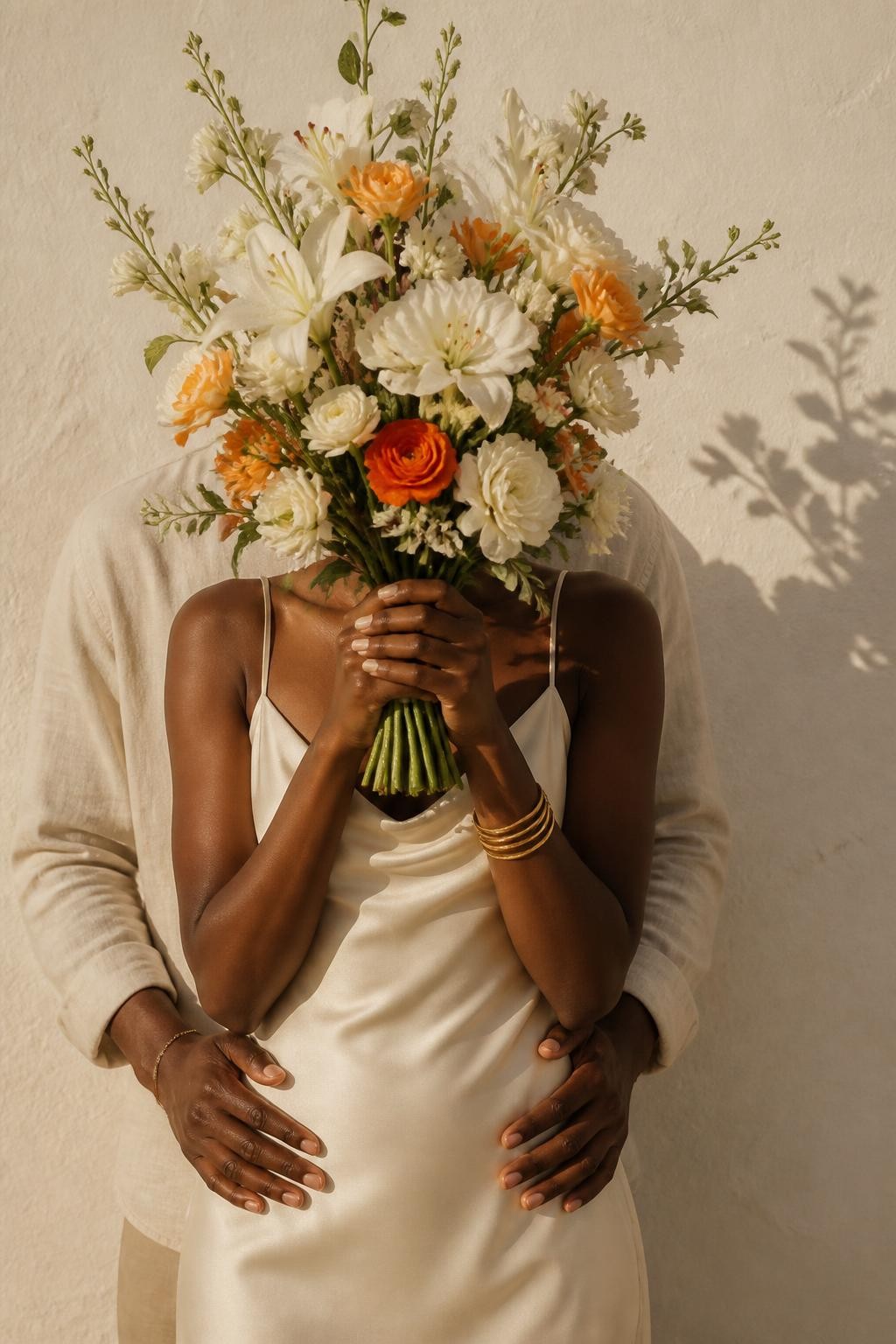 Image prompt: A stylish no-face couple portrait of an adult couple standing close against a textured cream wall, featuring a Black woman with deep brown skin wearing a soft ivory slip dress and gold bangles, holding a large bouquet of fresh white and orange flowers high enough to cover both faces completely, her partner standing behind her with his arms gently around her waist, only their torsos, hands, and the bouquet visible, no facial features showing, soft afternoon light, flower shadows on the wall, realistic petals, natural skin texture, elegant faceless romantic DSLR photography, modern editorial mood.